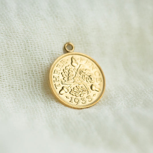 Gold coin pendant on a textured white fabric background showing acorns and oak leaves intertwined with Three Pence 1933 on the face of the coin