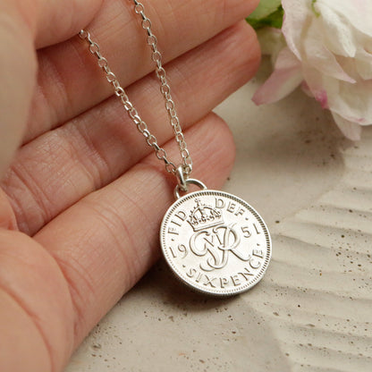 Silver sixpence necklace with a round pendant held by a hand, with flowers in the background.