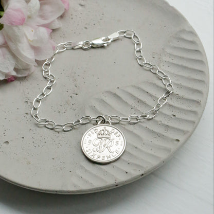 Silver sixpence British coin charm on an oval link bracelet with a coin charm on a concrete surface with flowers