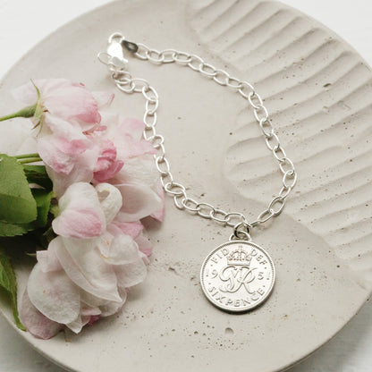 1951 silver sixpence British coin made into a charm with sterling silver and an oval chain bracelet. The bracelet sits on a background dish with blooms. 