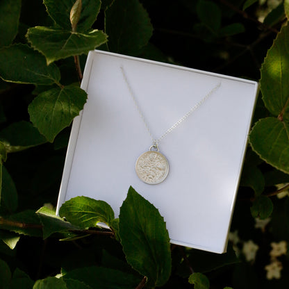 Silver necklace with a pendant on a white card, surrounded by green leaves.