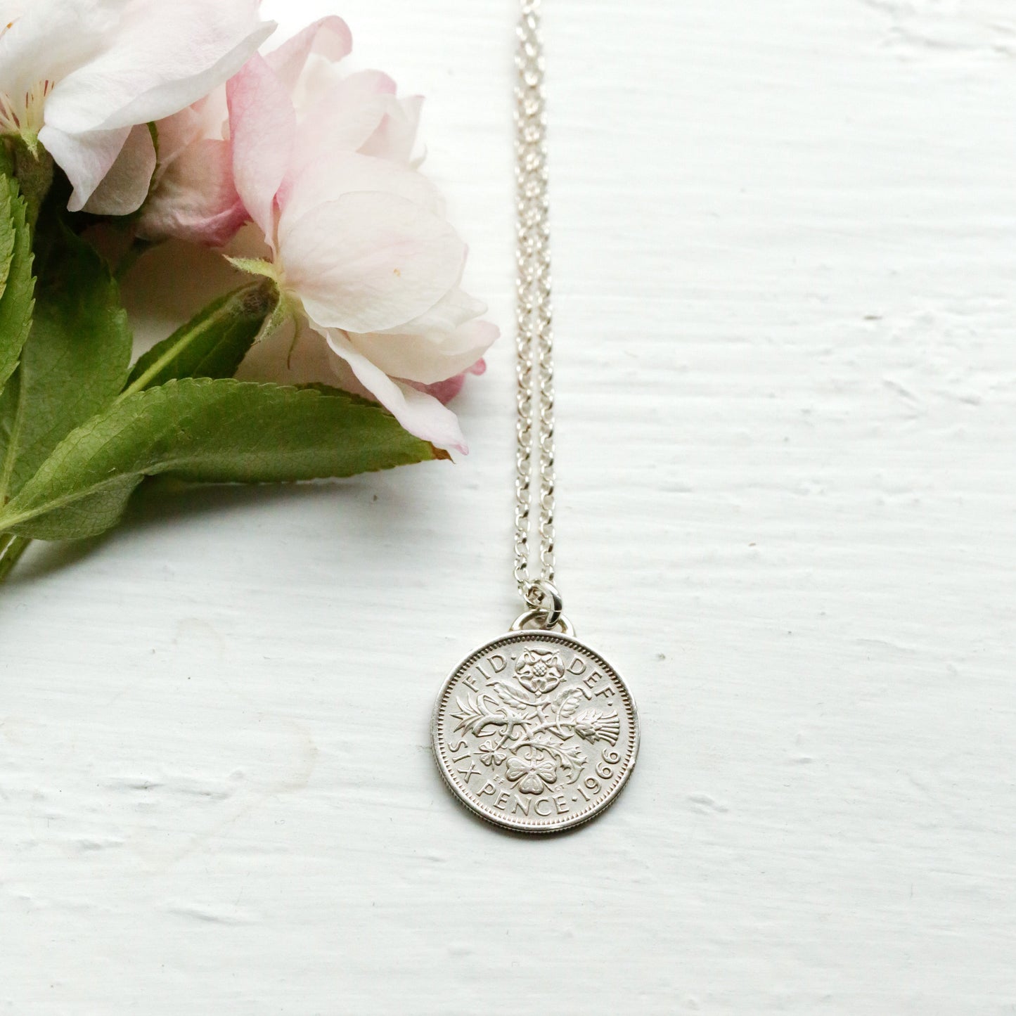 Silver sixpence necklace with a round pendant on a white surface with pink flowers. British coins made wearable jewellery.