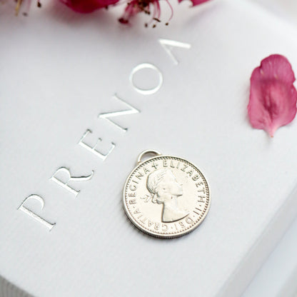 Silver coin pendant on a white box with 'Prenoa' branding, surrounded by pink flowers.