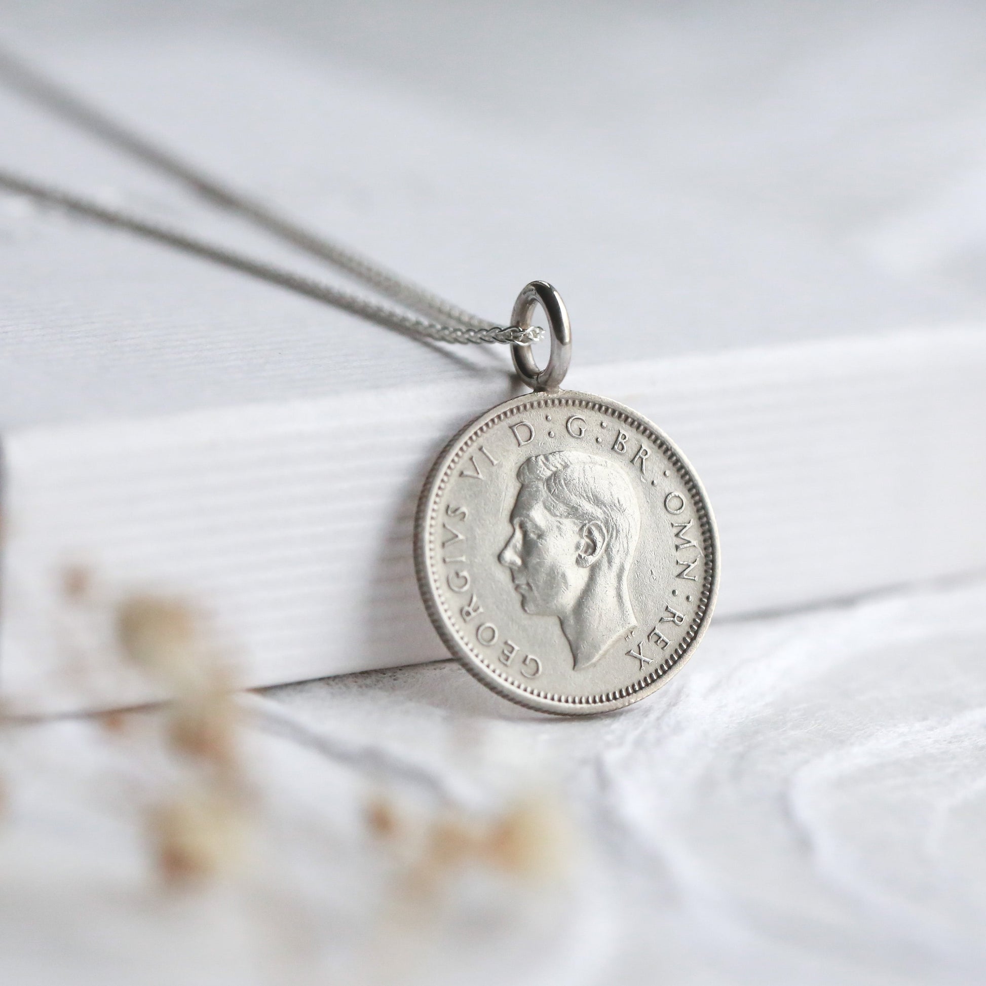 Silver sixpence pendant necklace on a white surface with dried flowers