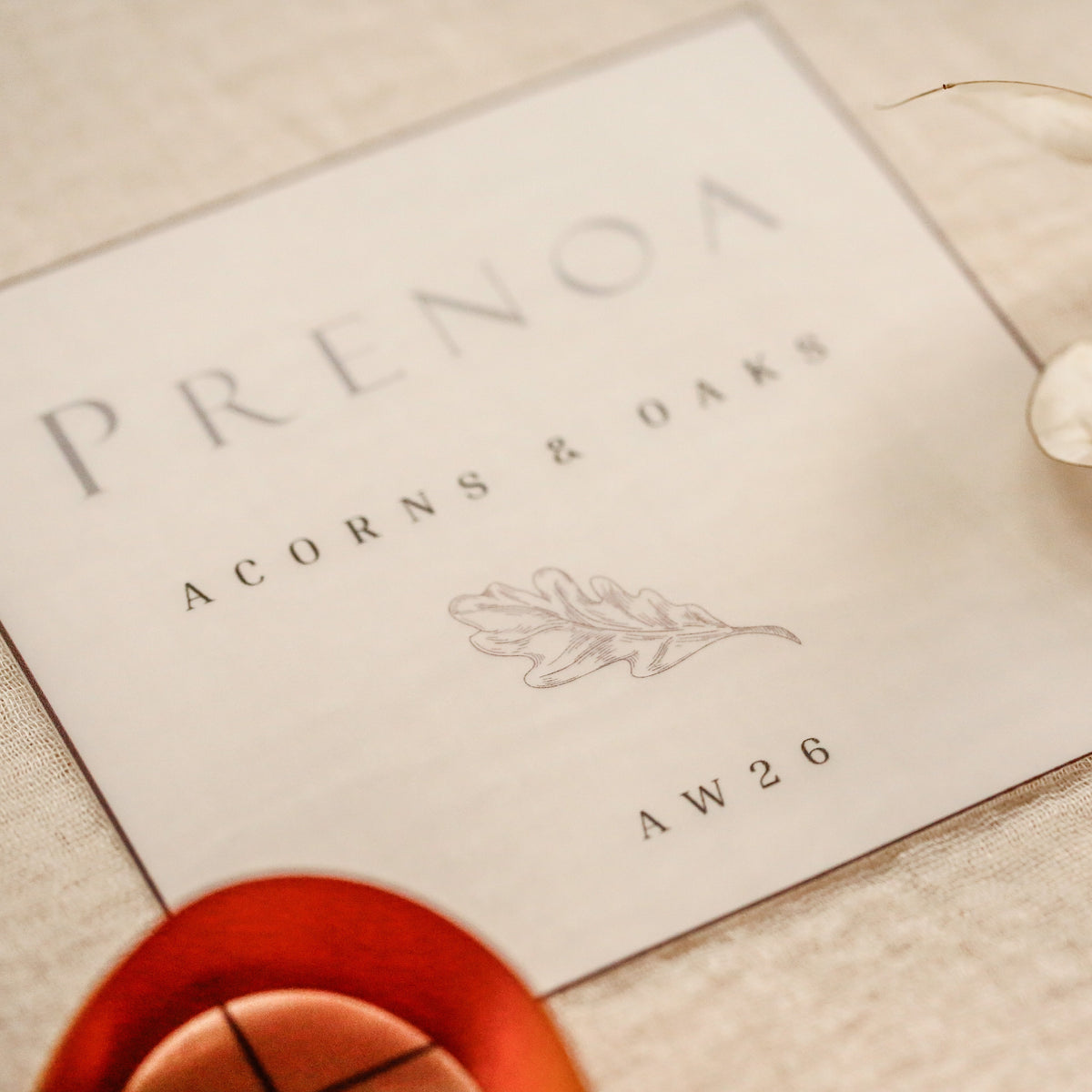 A square cream-colored card with the brand name PRENOA and the text 'ACORNS & OAKS' printed in gray, styled on a beige fabric surface with dried leaves and orange ribbon accents.