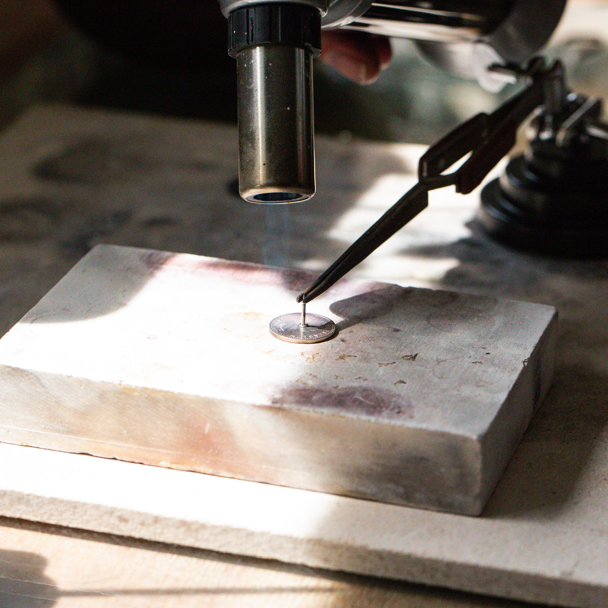 A real British sixpence lies on a oldering blog, with a sterling silver pin resting gently on top with the heat of a blow torch, soldering and permanently fusing metal to metal.