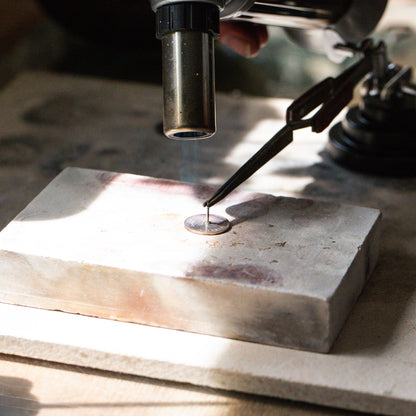 A real British sixpence lies on a oldering blog, with a sterling silver pin resting gently on top with the heat of a blow torch, soldering and permanently fusing metal to metal.