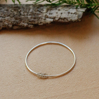 A delicate silver bangle bracelet with a small leaf-like wrap detail at the front, displayed on a textured beige surface with green pine branches and a piece of driftwood in the background.