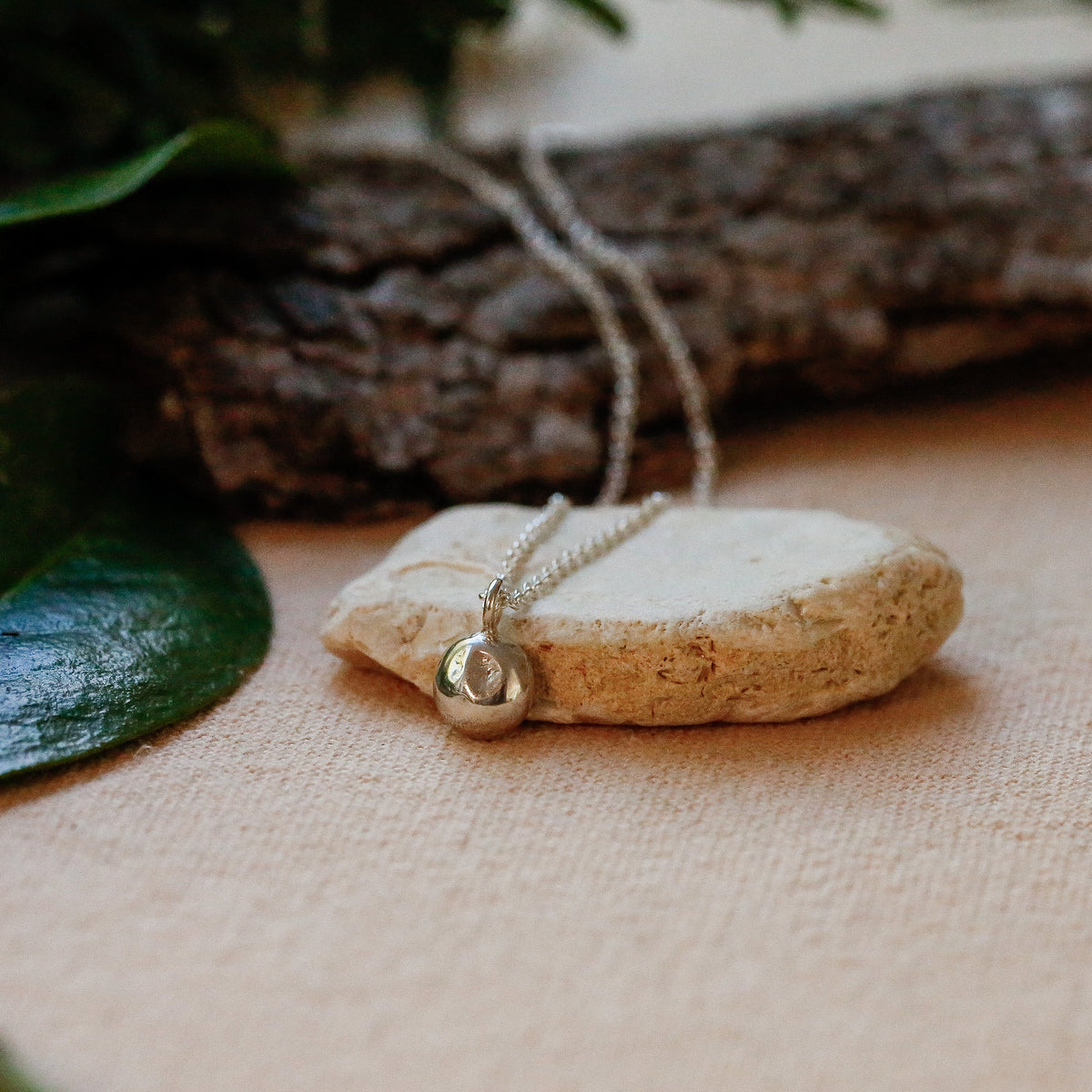 A natural sterling silver pendant necklace with a silver bail and chain, resting on a beige linen surface with a rustic wood branch and green leaves in the background.