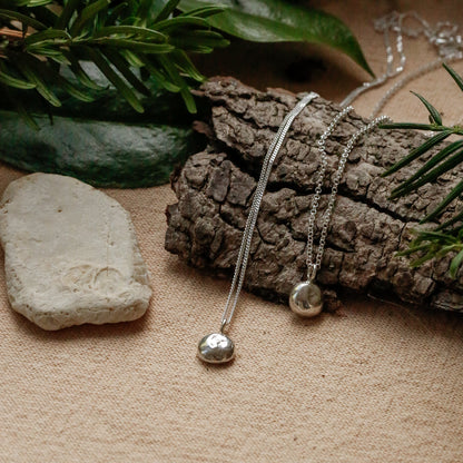 Two delicate silver necklaces with small spherical pendants are arranged on a textured beige surface with a piece of bark, green pine branches, and a small white stone.