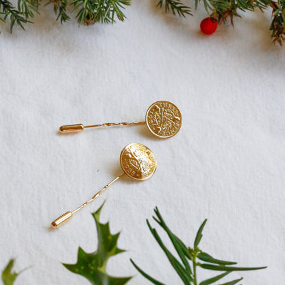 A pair of gold plated lapel pins with threepence coins and a twisted vermeil pin, arranged on a white textured paper background with green pine and holly branches as decorative props.