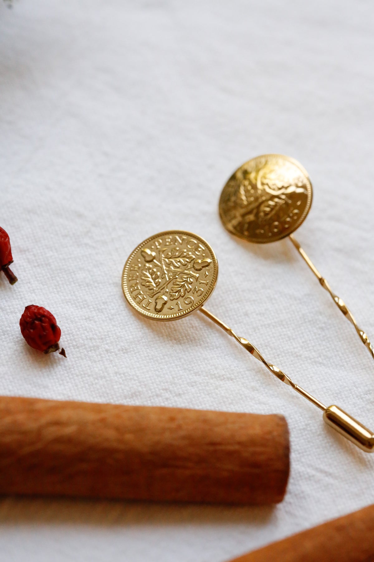 Two lapel pins with acorns and oak leaves design top and twisted wire stems, arranged on a white textured fabric with a cinnamon stick and small dried red berries as props.