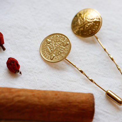 Two lapel pins with acorns and oak leaves design top and twisted wire stems, arranged on a white textured fabric with a cinnamon stick and small dried red berries as props.