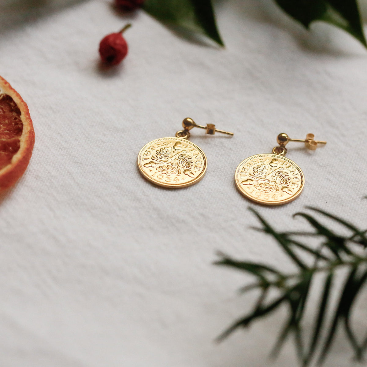 A pair of gold coin dangle stud earrings with a acorns and oak leaves design, displayed on a textured white fabric with dried botanical props.
