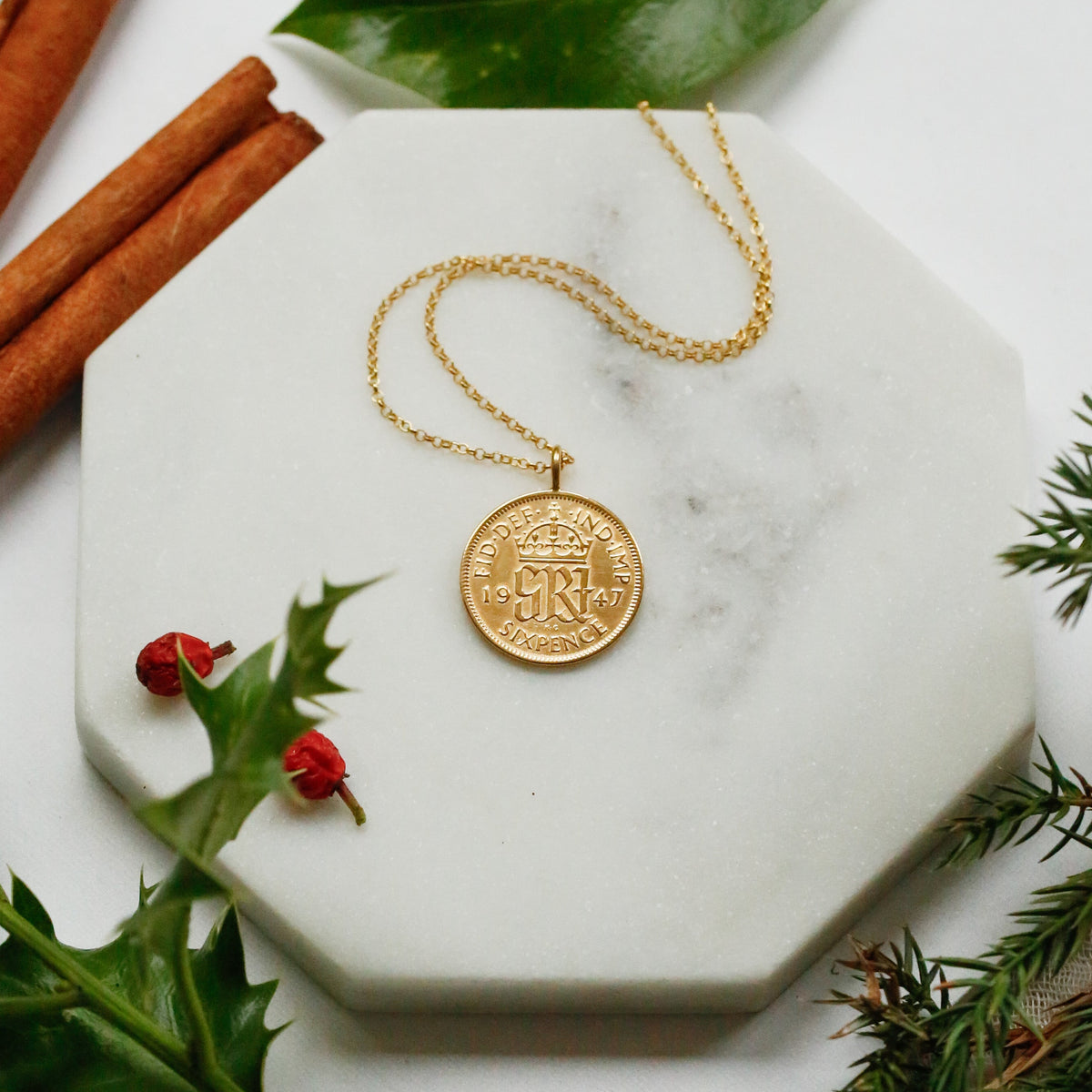 A gold sixpence coin pendant necklace with a crown emblem lies on a white marble hexagonal tray, styled with cinnamon sticks, green leaves, holly leaves, and pine branches on a white surface.