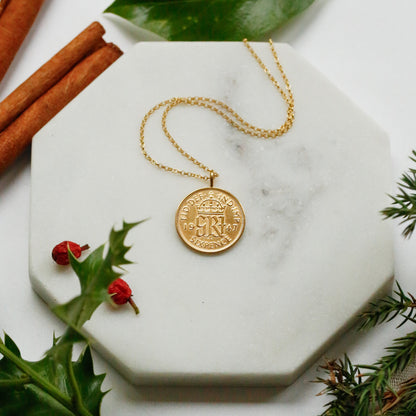 A gold sixpence coin pendant necklace with a crown emblem lies on a white marble hexagonal tray, styled with cinnamon sticks, green leaves, holly leaves, and pine branches on a white surface.