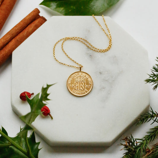 A gold sixpence coin pendant necklace with a crown emblem lies on a white marble hexagonal tray, styled with cinnamon sticks, green leaves, holly leaves, and pine branches on a white surface.