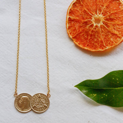 A delicate gold chain necklace with two small coin pendants, styled on a textured white surface alongside a dried orange slice and a green leaf.