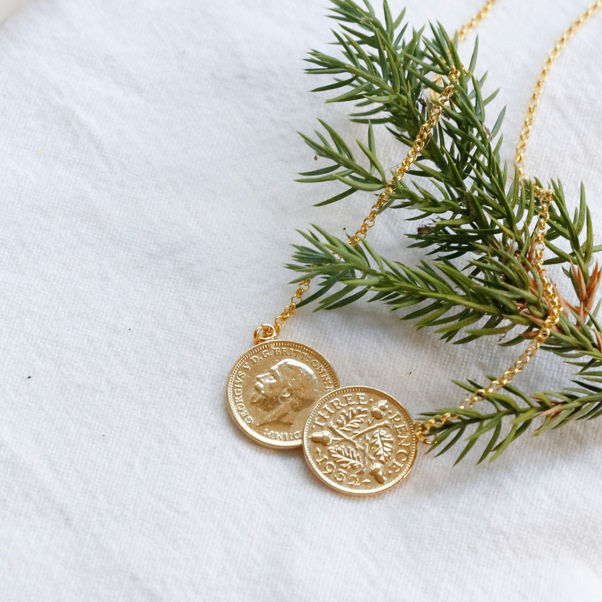 Gold double coin pendant necklace on a delicate gold chain, styled with a green pine branch on a textured white fabric background.