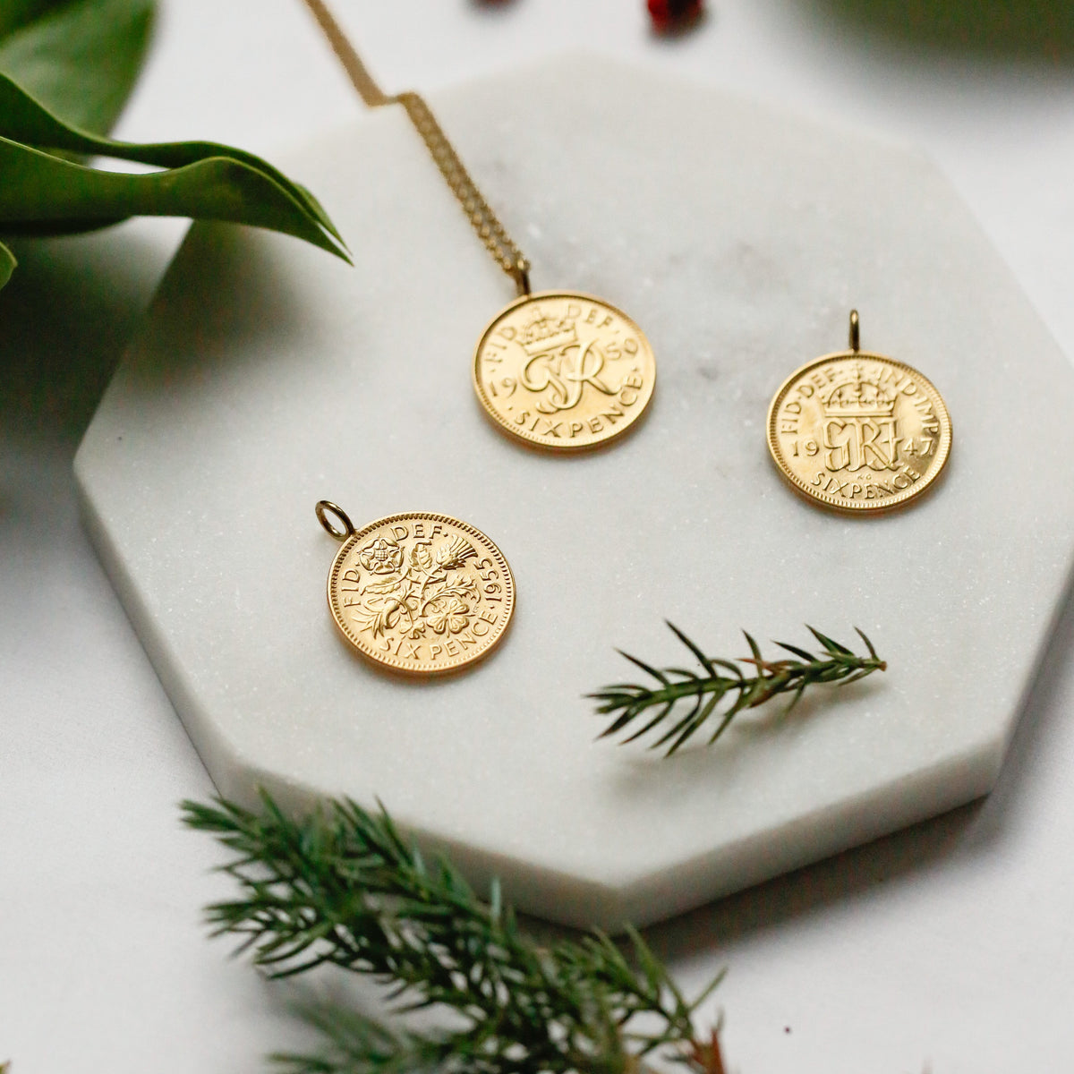 Three gold coin pendants using 22carat gold displayed on a white hexagonal marble tray, styled with green leaves and pine branches on a white background. Part of the British coin pendant collection gifts for him and her. 