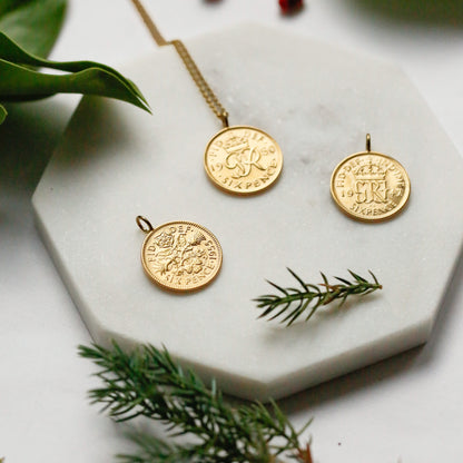 Three gold coin pendants using 22carat gold displayed on a white hexagonal marble tray, styled with green leaves and pine branches on a white background. Part of the British coin pendant collection gifts for him and her. 