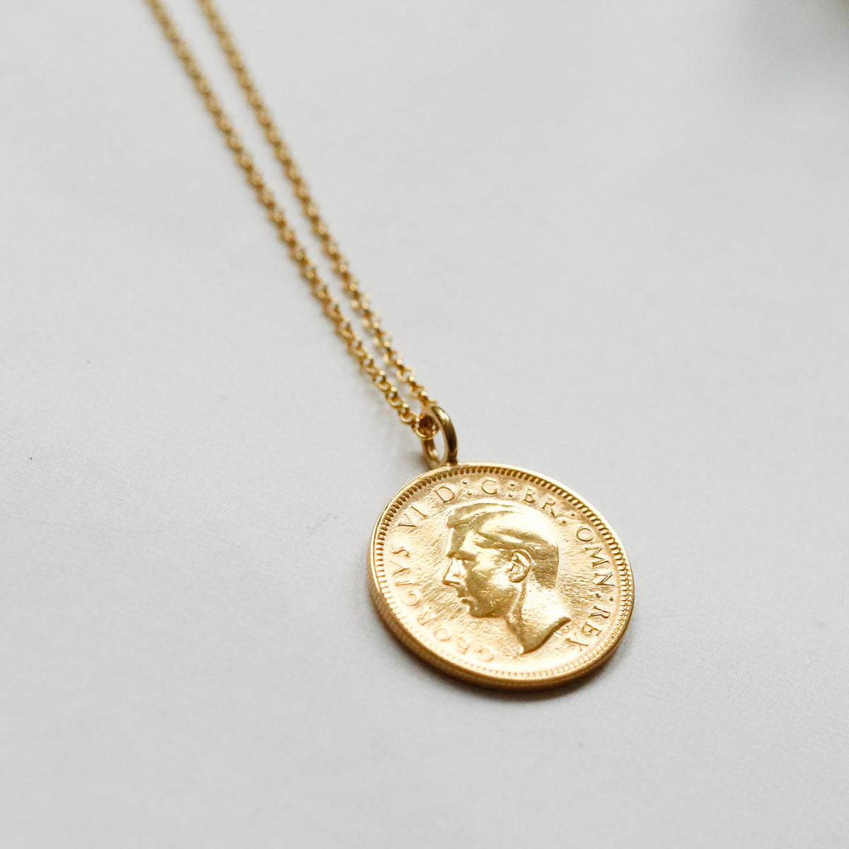 A gold coin pendant necklace featuring a profile portrait, suspended from a delicate gold chain against a light gray background.