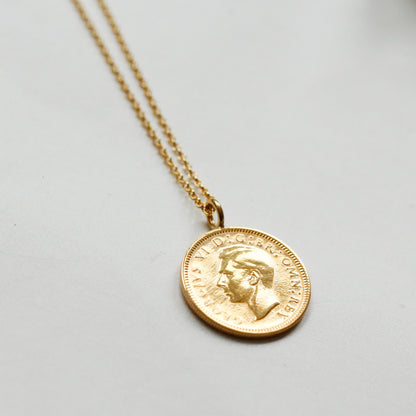 A gold coin pendant necklace featuring a profile portrait, suspended from a delicate gold chain against a light gray background.