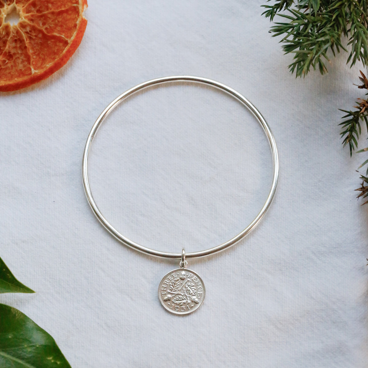 A thin silver bangle bracelet with a small circular coin pendant, displayed on a textured white surface with dried orange slices and green leaves as props.