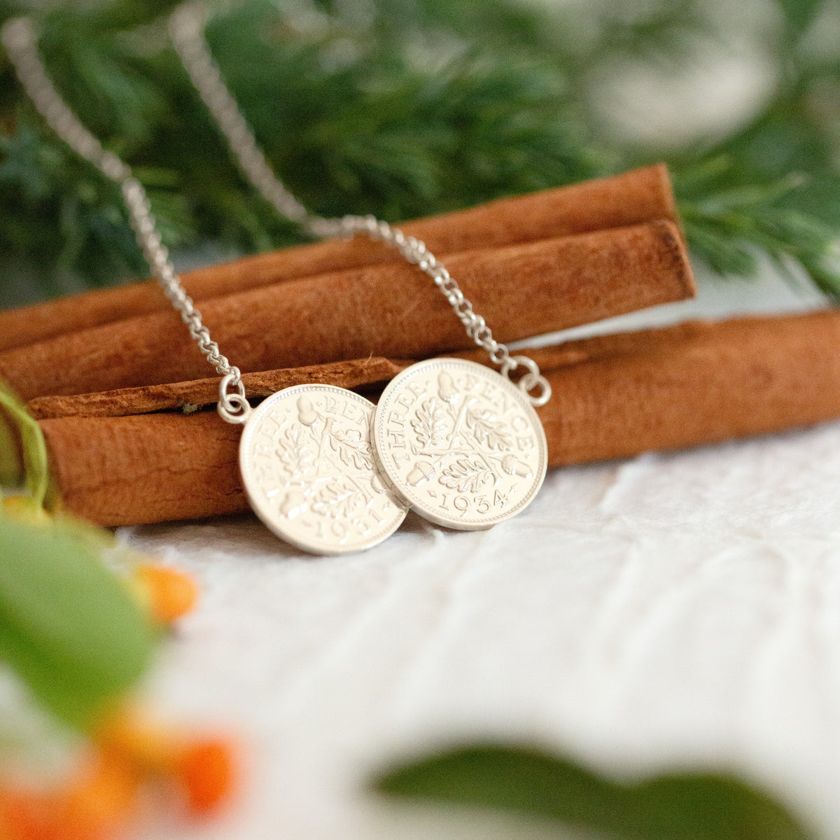 A pair of silver coin pendants on a delicate silver chain, styled on cinnamon sticks with green foliage and orange berries in the background.
