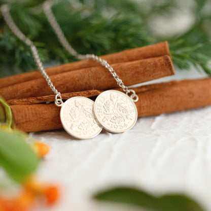 A pair of silver coin pendants on a delicate silver chain, styled on cinnamon sticks with green foliage and orange berries in the background.