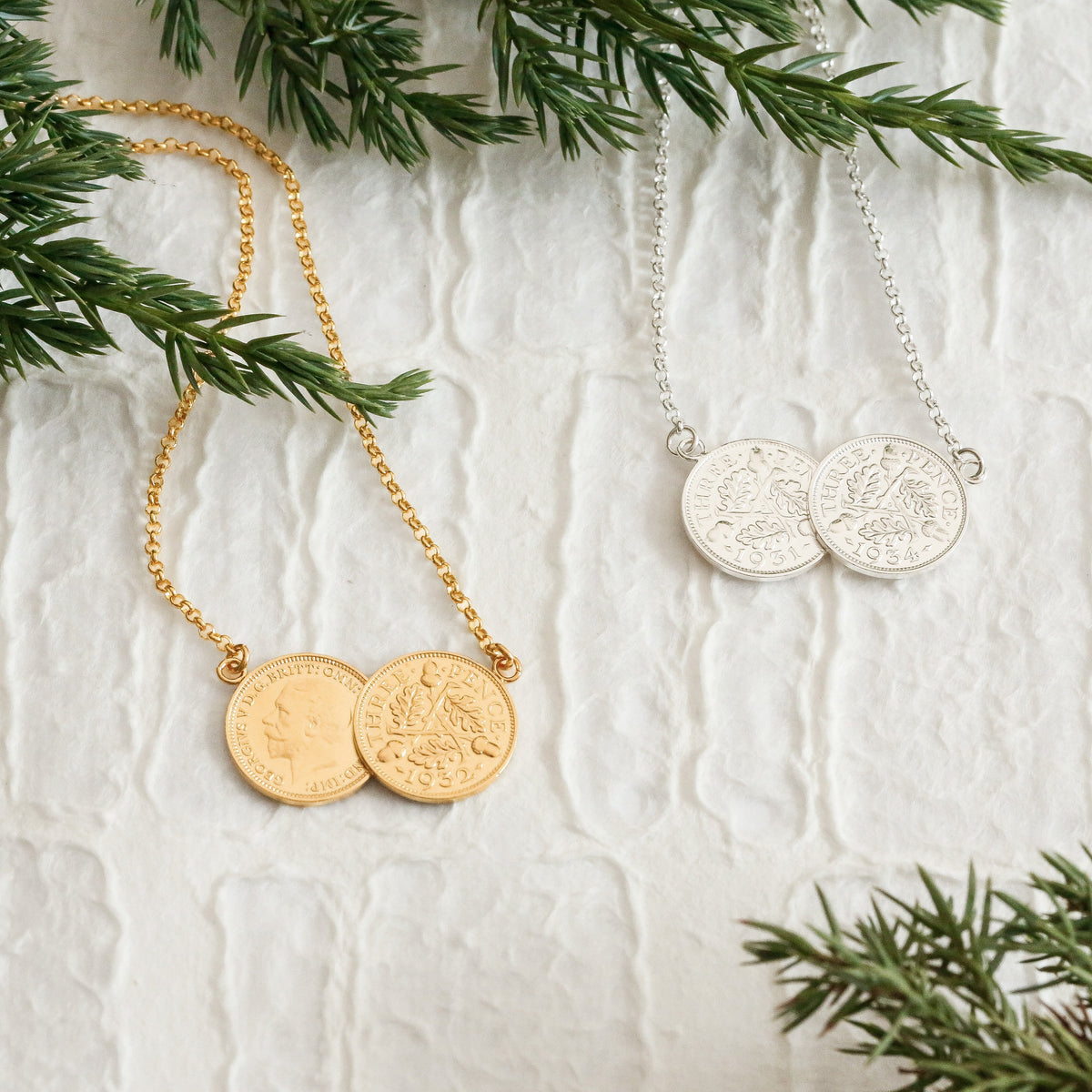 Silver and gold double coin necklaces made of two threepence coins, on a white paper surface with fir tree accents in the edges of the image.