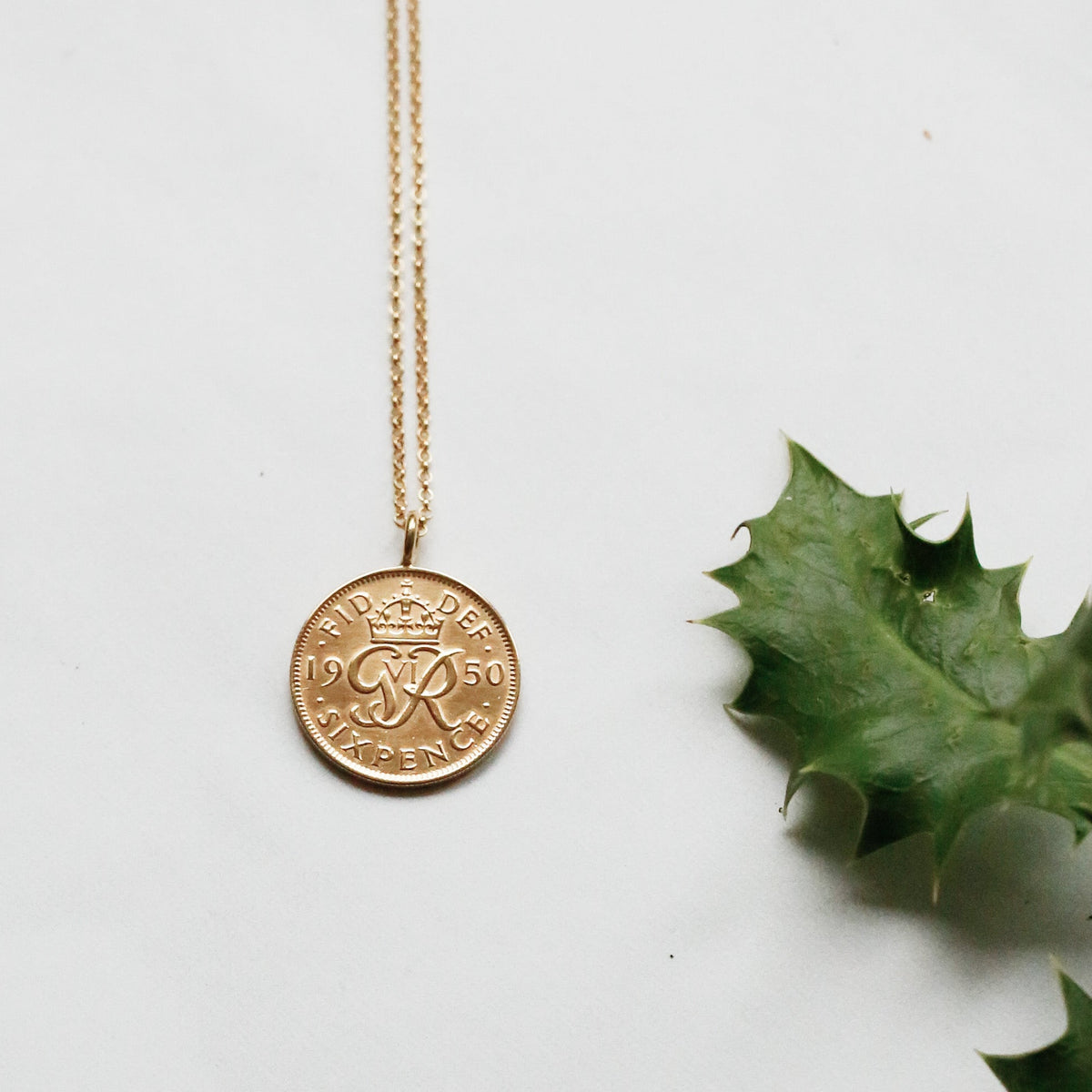 A gold sixpence pendant necklace with a circular coin-style charm displayed on a white surface with dried orange slices, a pinecone, and green holly leaves.