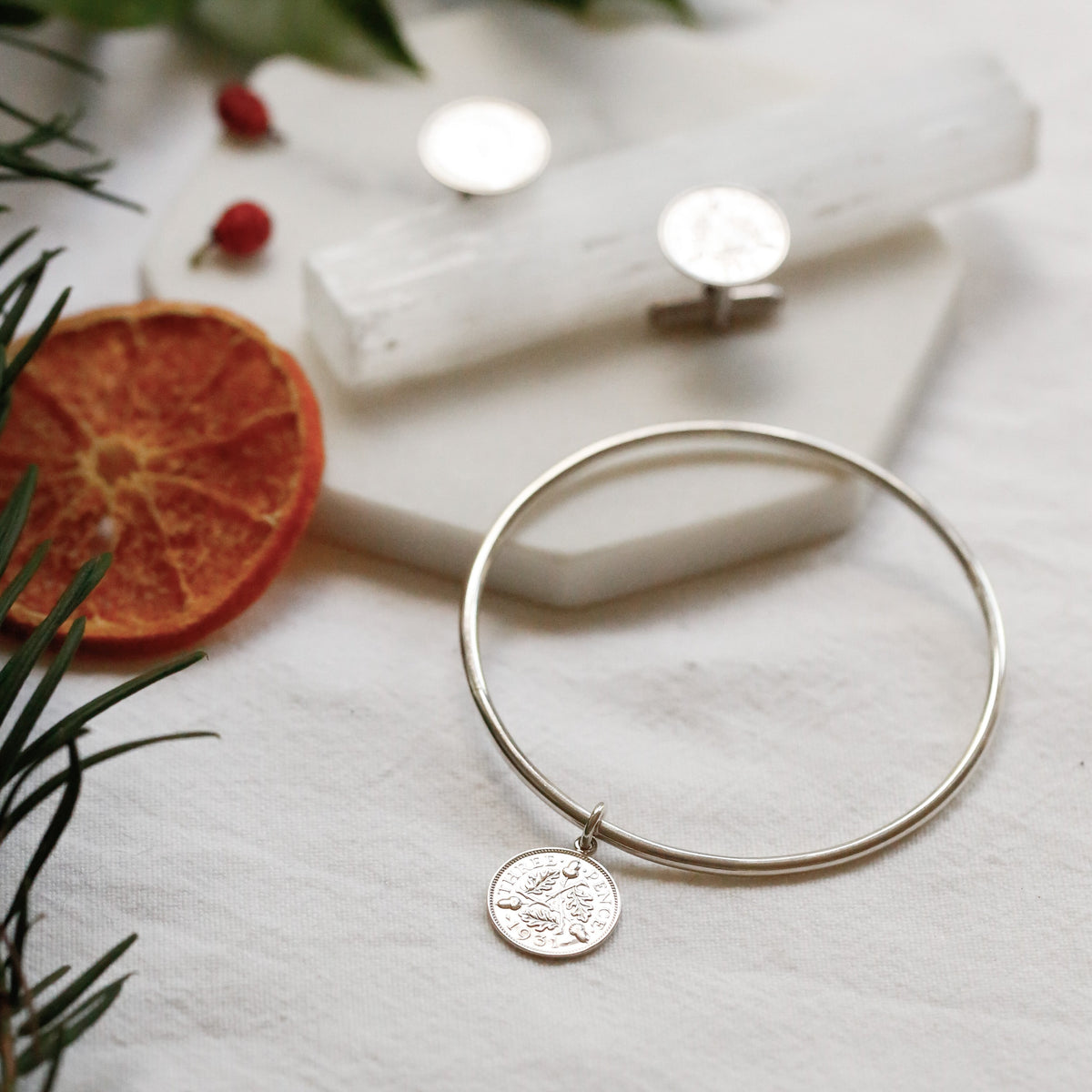 A thin silver bangle bracelet with a small round coin charm, styled on a white textured surface with a white crystal cluster, green leaves, and dried orange slices.