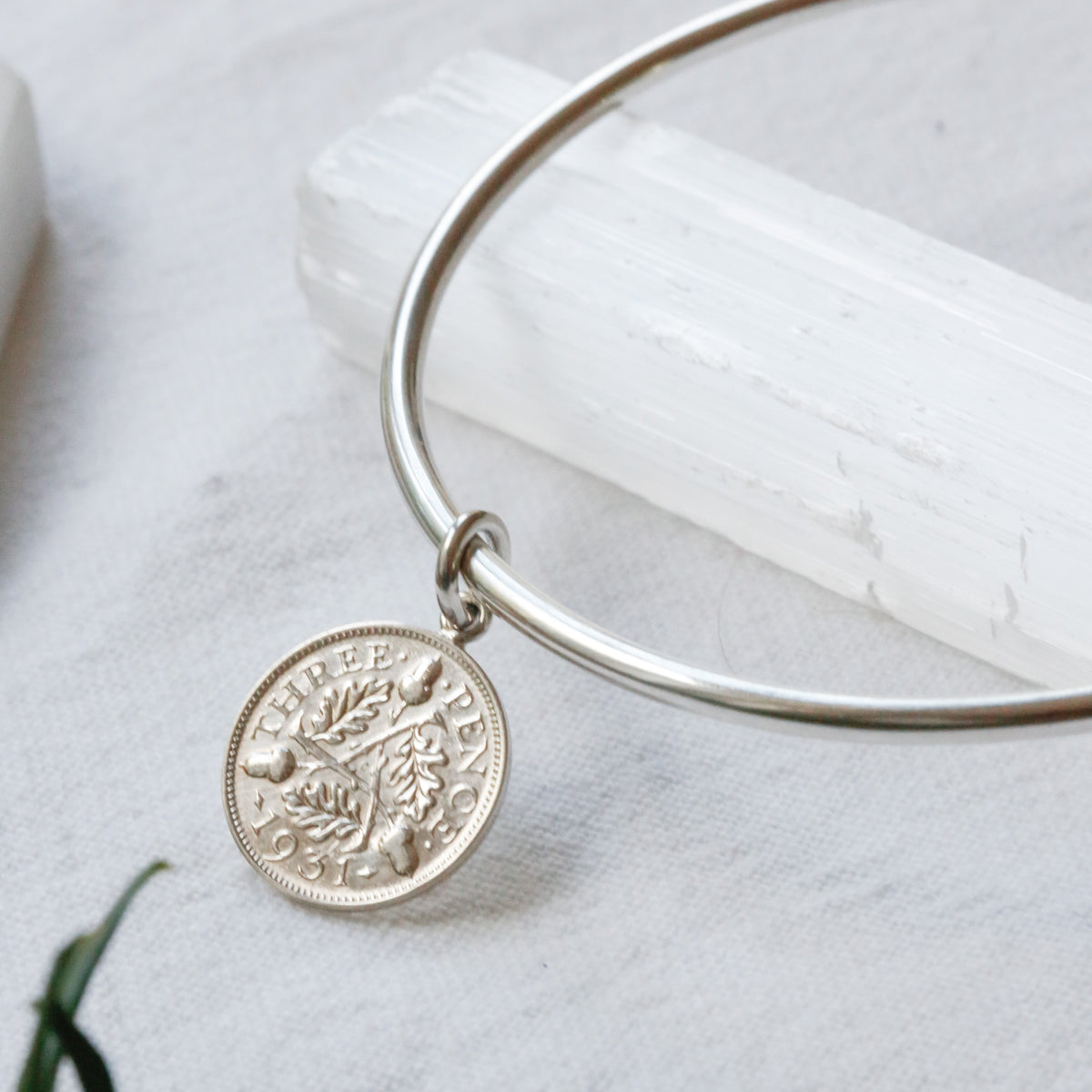 A silver bangle bracelet with a circular coin charm featuring a tree of life design, displayed on a white textured surface with a white crystal and green leaves.