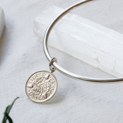 A silver bangle bracelet with a circular coin charm featuring a tree of life design, displayed on a white textured surface with a white crystal and green leaves.