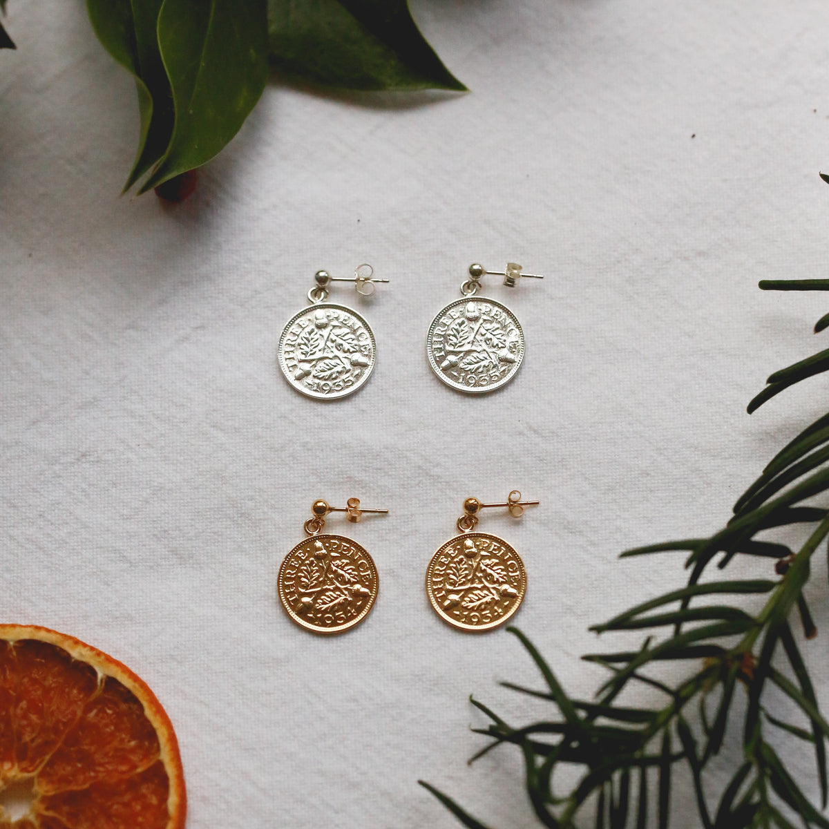 Two pairs of coin-style dangle earrings displayed on a white textured surface, styled with green leaves, a dried orange slice, and pine branches. One pair is silver-toned and the other is gold-toned, each featuring an embossed tree design.