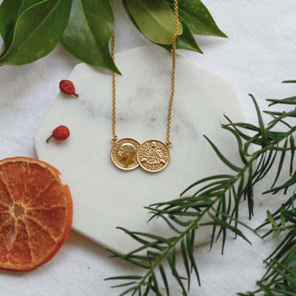 A gold double threepence British coin necklace with two round coins pendants is arranged on a white marble hexagonal board with green leaves, a dried orange slice, and small red berries on a white fabric background.