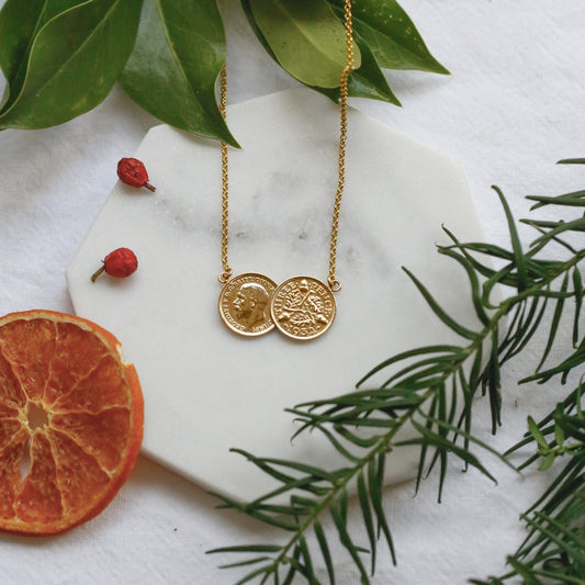 A gold double threepence British coin necklace with two round coins pendants is arranged on a white marble hexagonal board with green leaves, a dried orange slice, and small red berries on a white fabric background.