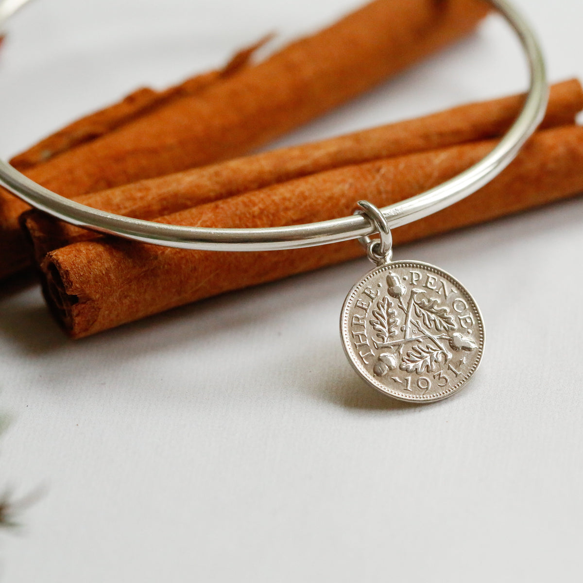 A silver bangle bracelet with a coin-style hanging charm, styled on a white surface with cinnamon sticks and small red berries.