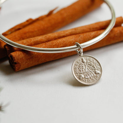 A silver bangle bracelet with a coin-style hanging charm, styled on a white surface with cinnamon sticks and small red berries.