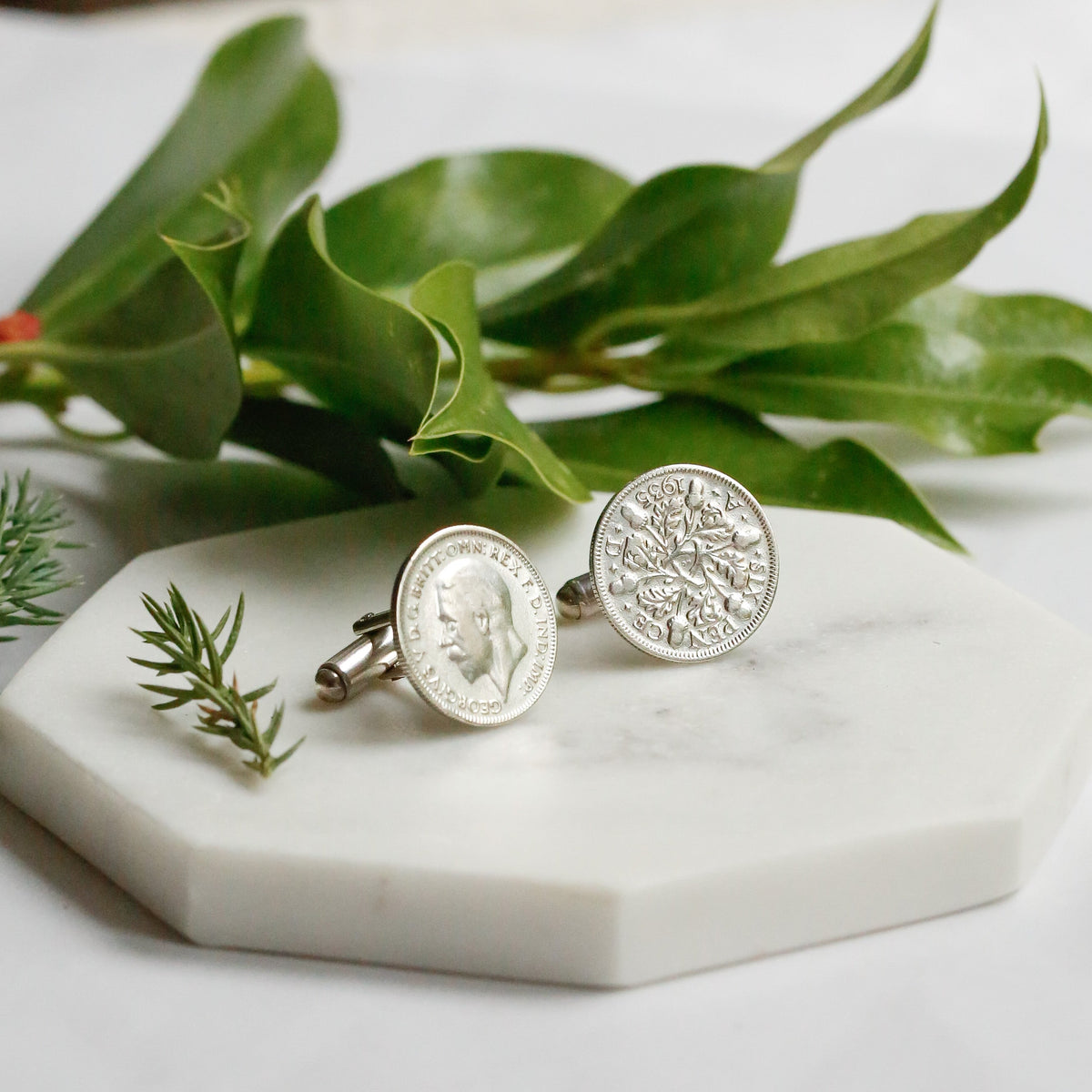 A pair of silver coin cufflinks with sterling silver spring backs sit on a marble plate with holly and berries in the background as a festive setting.