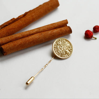 A sixpence lapel pin, plated in gold sits on styled on a white surface with cinnamon sticks, red berries, and green pine needles.