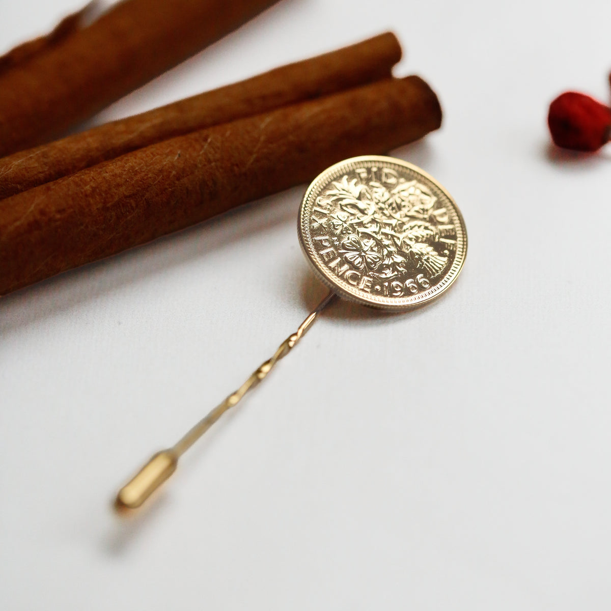 A gold sixpence lapel pin styled as a circular medallion pin, displayed on a clean white surface next to cinnamon sticks and a small red berry.