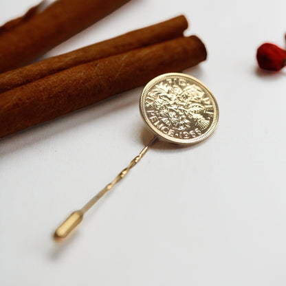 A gold sixpence lapel pin styled as a circular medallion pin, displayed on a clean white surface next to cinnamon sticks and a small red berry.