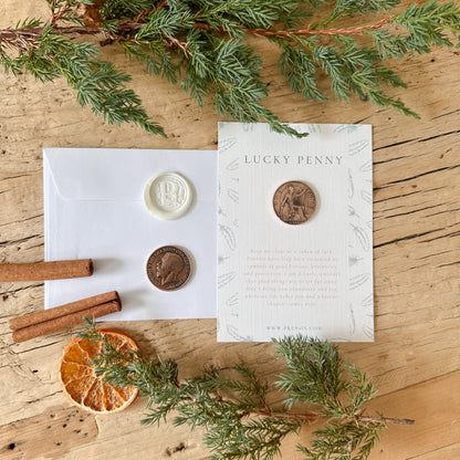 A flat-lay of a 'Lucky Penny' card and envelope set on a rustic wooden table, styled with green pine branches, cinnamon sticks, and a dried orange slice. The card features a copper-colored penny illustration and text, next to a white envelope with a white and bronze wax seal.