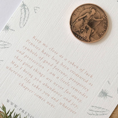A close-up of a white card with printed text and feather illustrations, featuring a bronze-colored penny placed on the card and a small green sprig at the corner, on a light surface.
