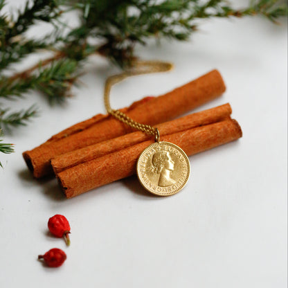 A QE2 sixpence necklace, plating in 22carat yellow gold and made into a pendant, is shown resting on some cinnamon with red berries in the foreground and a white background with fir leaves in the background.