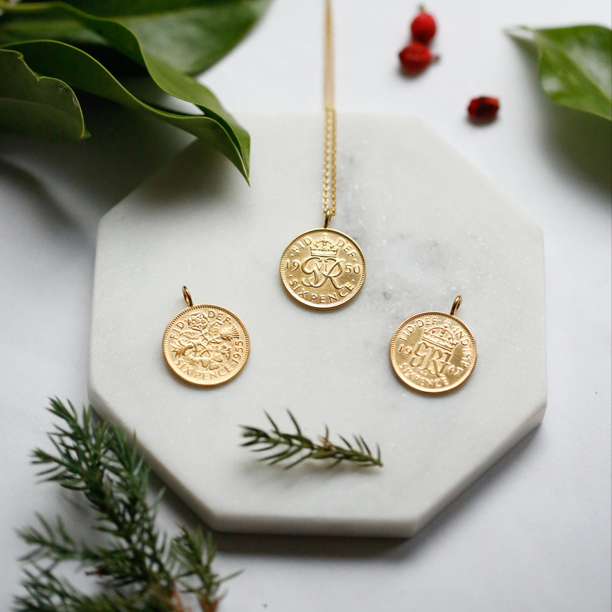 Three sixpences sit on a marble tile with foliage around them. The sixpence necklaces have been 22carat gold plated and make the perfect gift for his and her Christmas. 