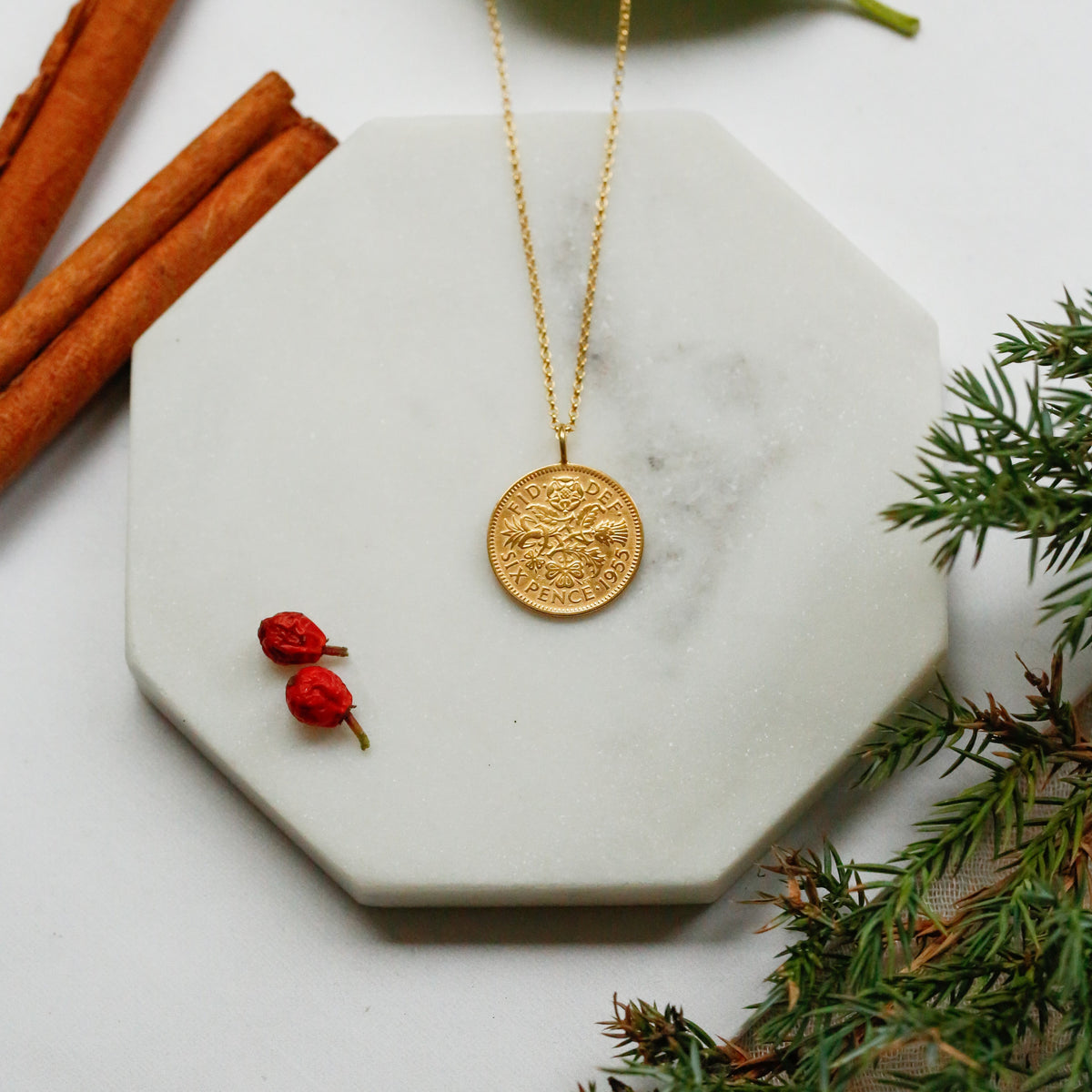 Queen Elizabeth II sixpence pendant gold plated in 22carat gold necklace with vermeil chain, sits on a marble coaster with cinnamon in the top left of the image with red berries and festive fir detailing. The ideal Christmas gift rich in tradition made in Britain. 