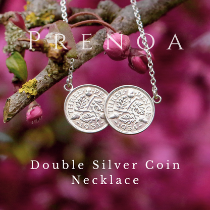 Double silver coin necklace on a branch with pink flowers and leaves, branded 'Prenoa'.
